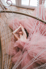 young girl with short black hair in pink tulle dress is lying under crystal chandelier near white window on the wooden floor background at sunny day, camera from top. fashion concept, free space