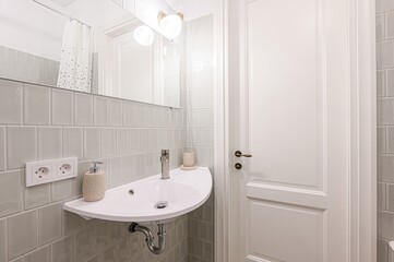 bright bathroom features a half-circle sink, chrome faucet, soap dispenser, and electrical outlets. Light grey tiles cover the walls. A white door with a brass handle is visible