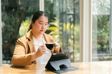 Asian Businesswoman Young woman enjoying coffee while using a tablet in a bright, plant-filled...
