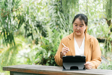 Fototapeta premium Asian Businesswoman A woman writes on a tablet in a lush green outdoor setting, exuding a sense of focus and productivity amidst nature. Female Digital nomad lifestyle