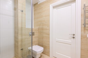 clean, modern bathroom with light wood-like tile, a white toilet, glass shower partition, and a white door