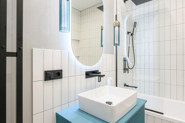 modern bathroom with white vertical tiles, a square sink on a blue cabinet, a round mirror with LED lighting, and black fixtures