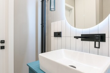 Modern bathroom detail with a white rectangular sink, blue cabinet, round mirror with light, and black fixtures against white tiled walls