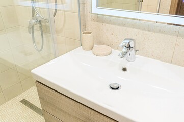 Fototapeta premium clean, well-lit bathroom sink area is shown with a white basin, chrome faucet, and beige accessories. A shower is visible in the background.