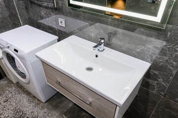modern bathroom scene features a white sink vanity, washing machine, and gray marble-like tiles. A lighted mirror hangs above.