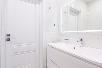 bright, modern bathroom featuring white marble walls, a sleek white vanity, a chrome faucet, and an illuminated mirror. A closed white door is seen on the left.