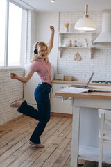Portrait of smiling pretty young teenager dancing on the kitchen, wearing headset opposite laptop