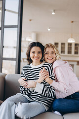 Obraz premium Two amazing girls sitting on the sofa, hugging one another, smiling lovely, looking at the camera