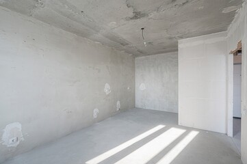 Bare, light-filled room in renovation with exposed concrete ceiling, plastered walls, and smooth...