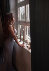 Fototapeta premium young brunette girl with long hair in blue silk dress is standing in profile near huge white window with old retro candles in vintage style interior background. lifestyle concept, free space