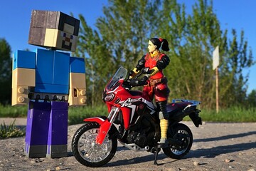 Fototapeta premium GI JOE action figure of female intelligence agent Jinx, version 2004 by Hasbro, standing on red dual sport motorbike Honda Africa Twin, meeting LEGO Minecraft figure of Steve on spring asphalt road. 