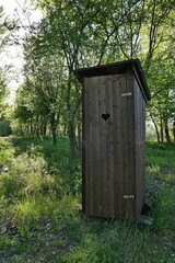 Narrow wooden latrine with typical hearth shaped small opening on doors, placed on pathway next to broadleaf trees surrounding fish pond. Spring daylight sunshine. 