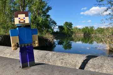 Naklejka premium LEGO Minecraft action figure of smiling mainc character Steve walking on concrete pipeline bridge across large spring fish pond, broadleaf trees and reed plants visible on bank. Blue skies with clouds