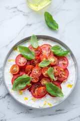 Stracciatella or stretched curd cheese with red cherry tomatoes and fresh basil, vertical shot on a white marble background, high angle view