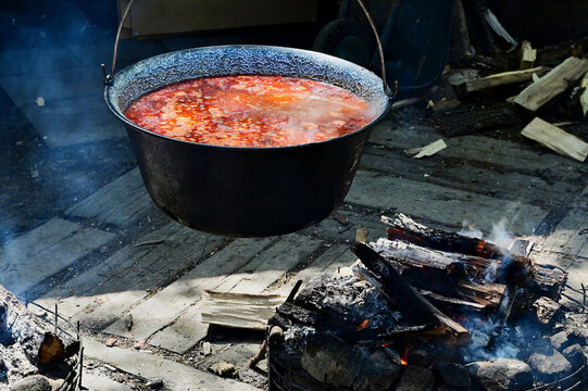 making goulash soup on open fire in large suspended kettle. red, spicy food. food and gastronomy concept. split firewood. smoke rising. closeup view. selective focus. blurred background. 