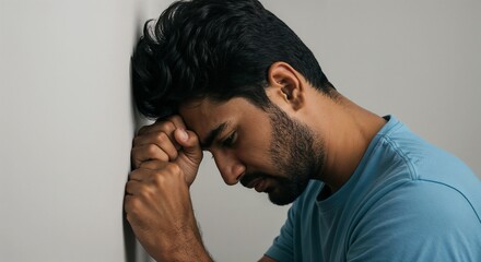 young South Asian man leaning forehead on light gray wall, fists clenched, emotional breakdown moment