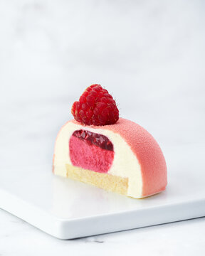 Individual raspberry and white chocolate mousse cake, sliced in half. Velvet texture, visible layers and fresh raspberry topping. White plate over marble background with copy space.