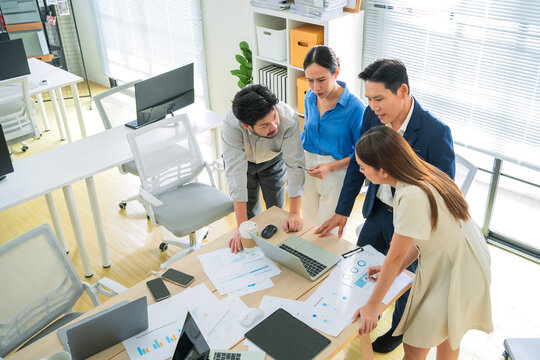 Standing business professionals in a strategy meeting, reviewing financial charts and laptop reports, brainstorming ideas for success and performance growth