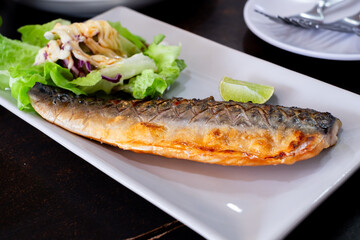 Grilled mackerel served on a white plate with fresh green salad, topped with creamy dressing and a slice of lime. A healthy and delicious seafood dish perfect for food content