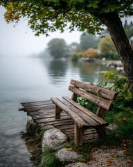 Fototapeta premium Rustic bench on dock by calm lake on a foggy day, tree overhead. Serene scene