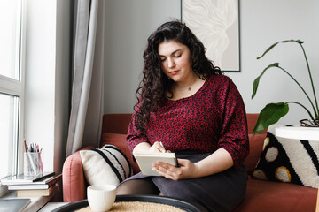 Portrait of young concentrated plus size woman writing in notebook sitting at cafe table on couch next to window with white mug of coffee, focused on making to-do list or noting down goals