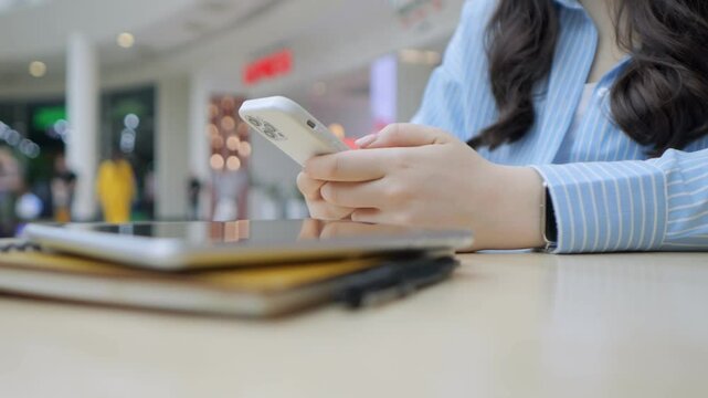 Woman is holding a cell phone and looking at it. She is sitting at a table with a stack of books and a laptop. The scene suggests that she might be working or studying, as she has multiple devices