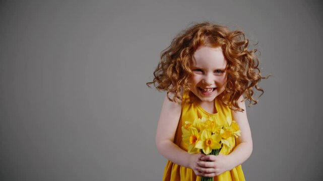 Smiling curly-haired girl in yellow dress holding daffodils against neutral background


