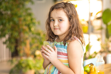 Fototapeta premium Cute little kid girl have breakfast holding a glass of green smoothie at home in the kitchen. Healthy food.