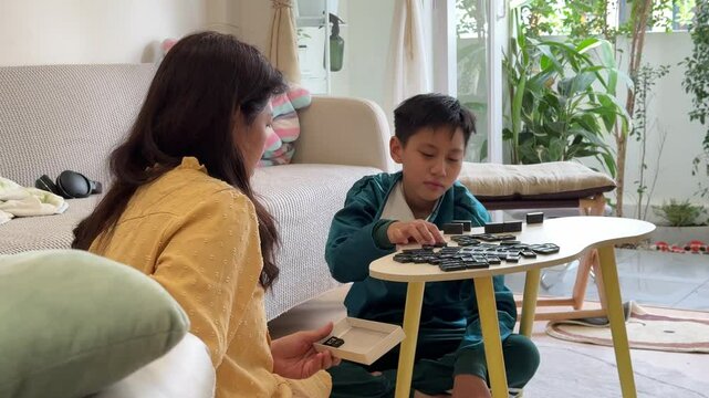The Southeast Asian boy is playing a domino game with his mother in the living room.