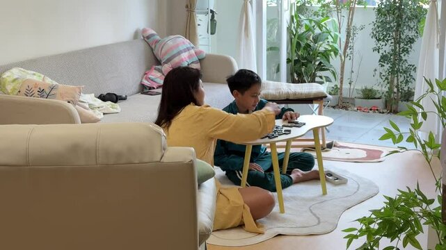 The Southeast Asian boy is playing a domino game with his mother in the living room.