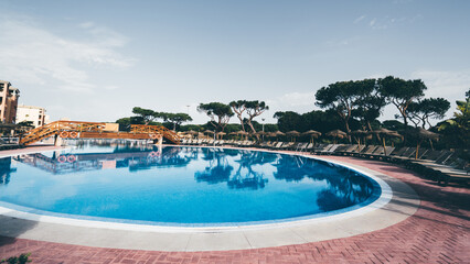 Serene view of a large circular outdoor swimming pool with clear blue water, surrounded by empty lounge chairs and palm trees, featuring a decorative wooden bridge and tranquil resort atmosphere