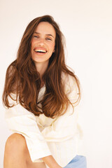 Fototapeta premium Portrait of a cheerful young woman with long brown hair, wearing a loose white shirt, sitting relaxed and smiling warmly. Natural light studio shot. Happiness, beauty, wellness, lifestyle.