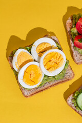 Delicious healthy bread toasts with fresh avocado, boiled eggs, cherry tomato and cucumber placed on bright yellow background