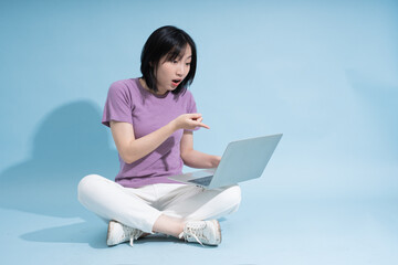 Young Asian woman shows surprise while using a laptop on a light blue background, expressing...
