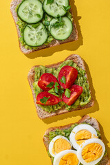 Delicious healthy bread toasts with fresh avocado, boiled eggs, cherry tomato and cucumber placed on bright yellow background