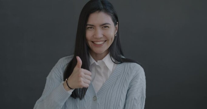 Slow motion portrait of joyful Asian lady showing thumbs-up hand gesture laughing looking at camera on black color background.