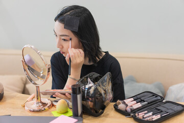 Young Asian woman applying makeup at a vanity with brushes and tools in a cozy setting during the...