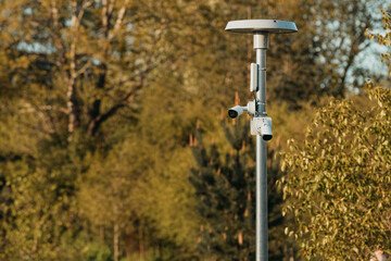 Two white surveillance cameras mounted on a metal pole with a circular antenna, set against a blurred background of trees and foliage.
