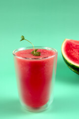 A glass of watermelon juice with a sprig of mint on top. A slice of watermelon is on the table next to the glass