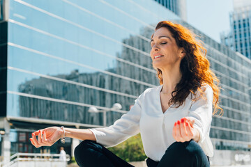 Professional businesswoman meditating in lotus position near sleek skyscrapers, demonstrating workplace wellness and stress management strategies in contemporary urban setting