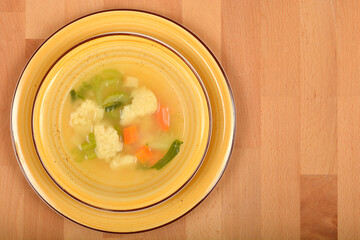 A close-up shows a yellow bowl filled with clear vegetable soup containing semolina dumplings, carrots, and green herbs