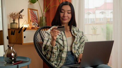 Happy Asian Korean woman female indoor holding bank credit card laptop computer online order...