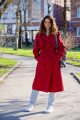 Obraz premium Stylish female student wearing a vibrant red trench coat and white sneakers enjoys a sunny spring day, strolling through a park while carrying a backpack and radiating confidence