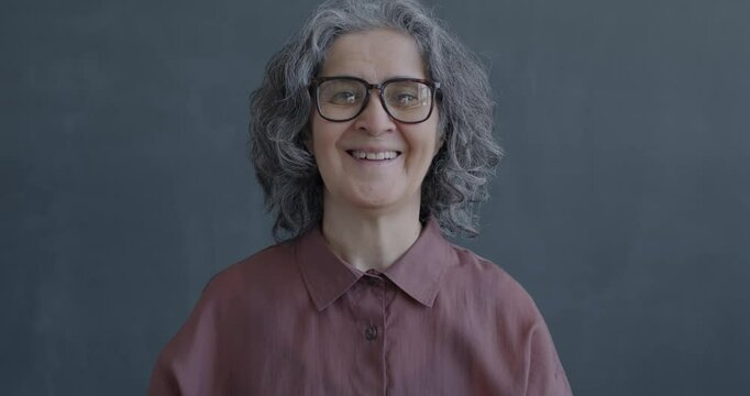 Slow motion portrait of happy senior woman showing OK hand gesture smiling on gray color background. People and positive emotion concept.
