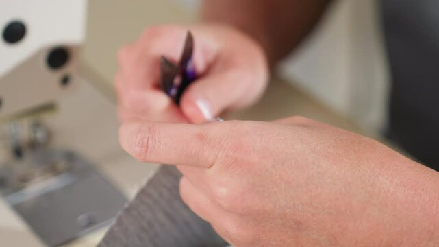 Close up designer hands cutting extra loose threads from fabric edge with small precision scissors focusing on removing imperfections while sewing near machine in brightly lit creative workshop