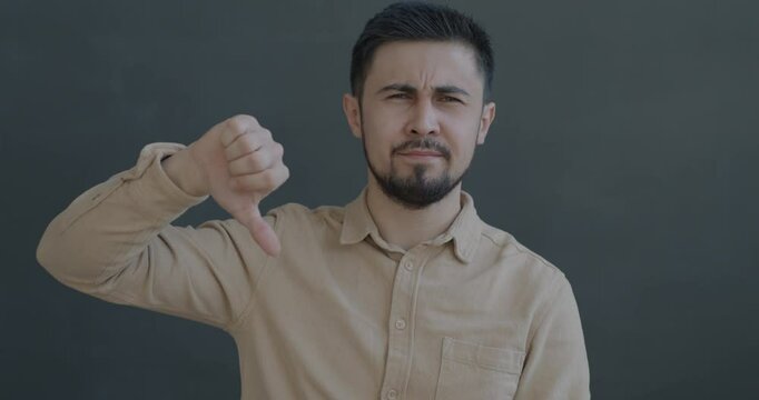 Slow motion portrait of unhappy biracial man showing thumbs-down hand gesture looking at camera on black color background