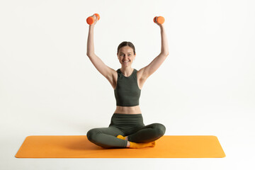 A woman sits cross-legged on an orange yoga mat, lifting two orange dumbbells above her head. She...