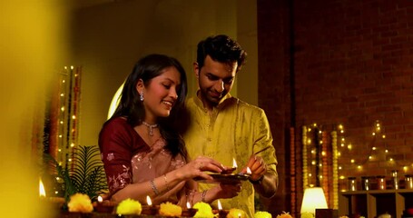 Indian couple wishing Happy Diwali while placing lighting decorative oil lamp diya on wooden railing, looking at camera smiling joyfully in traditional attire during Deepavali Hindu festival night