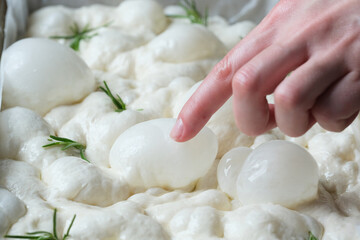 A hand gently presses into fluffy focaccia dough with visible bubbles and sprigs of fresh rosemary,...