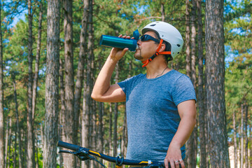 Cycling Fitness: An adult sport enthusiast in sportswear paused riding to drink water, amidst an urban park in surrounding by pine trees. This photo is part of a series themed by outdoor activities.